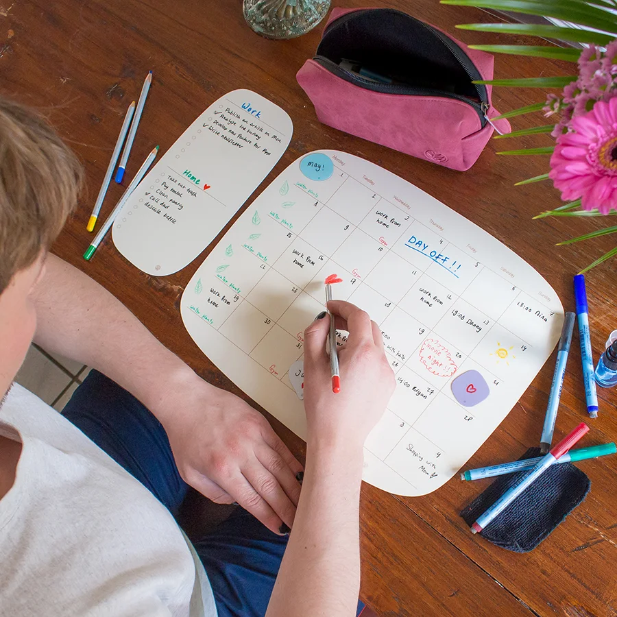 ein Mann schreibt auf einem Tisch mit einem organisch gestalteten Monatsplaner, neben dem Monatsplaner liegt ein Klebezettel, außerdem liegen Stifte auf dem Tisch verstreut