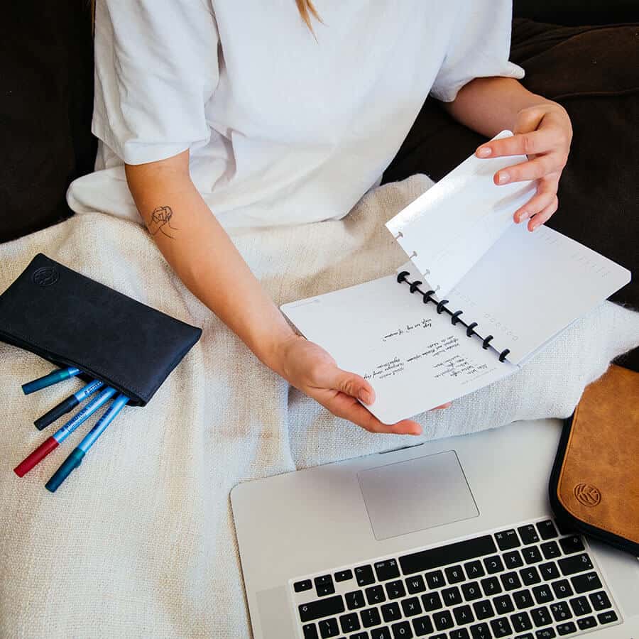 Das Modell blättert in einem Whiteboard-Notizbuch, rechts von ihr befindet sich ein Federmäppchen, links von ihr ein cognacfarbenes Arbeitsbuch und vor ihr ein Laptop
