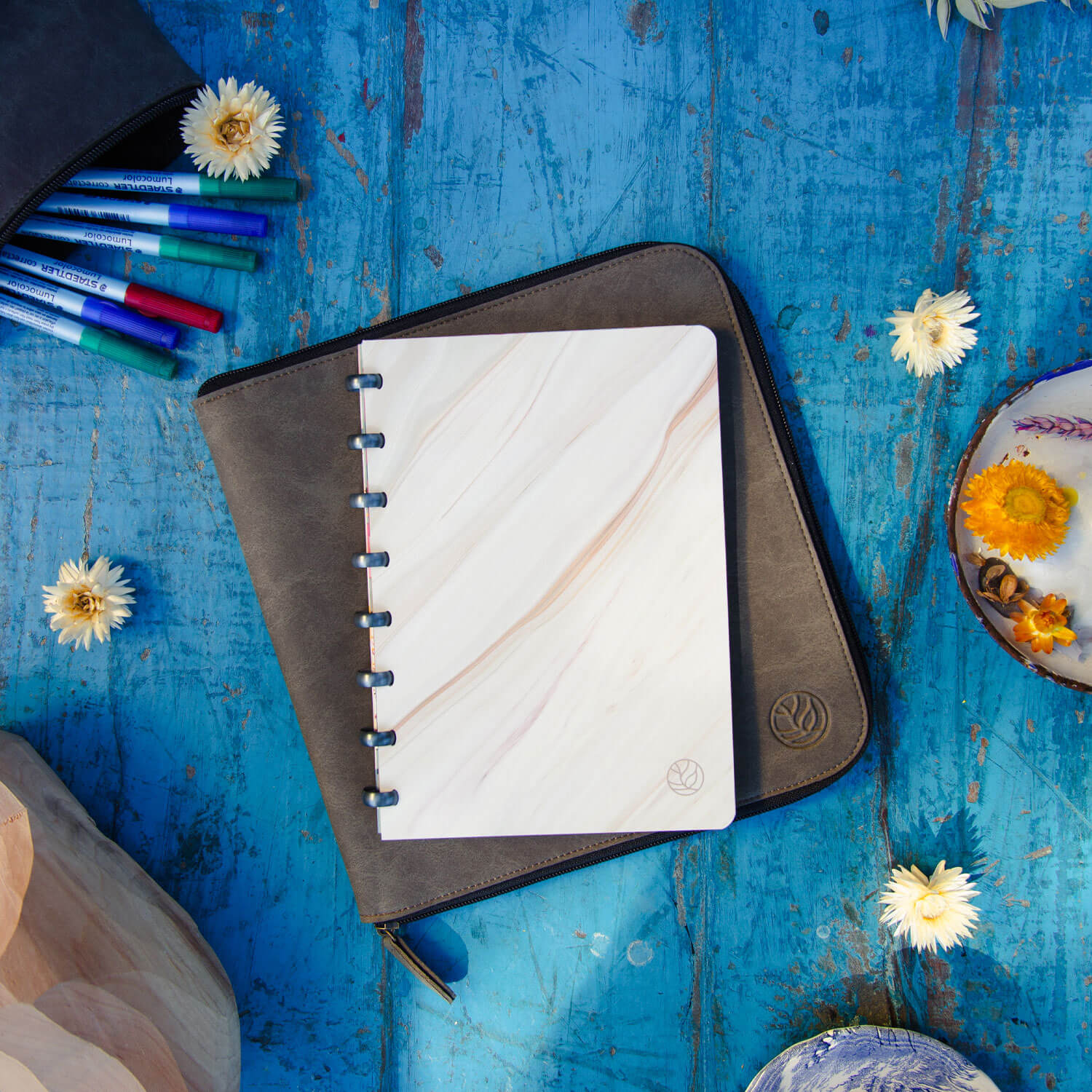 ein Whiteboard-Notizbuch mit fließendem Marmoreinband und raumgrauen Ringen, das auf einer Arbeitsmappe liegt, die wiederum auf einem antiken blauen Holztisch liegt